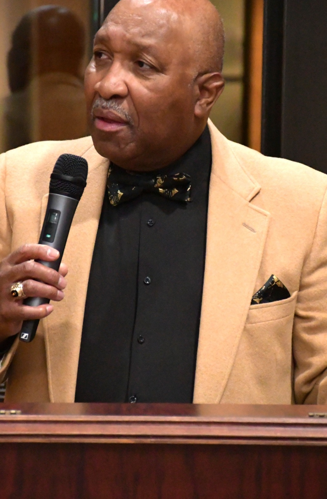 A speaker in a tan jacket speaking into a microphone at the alumni celebration.