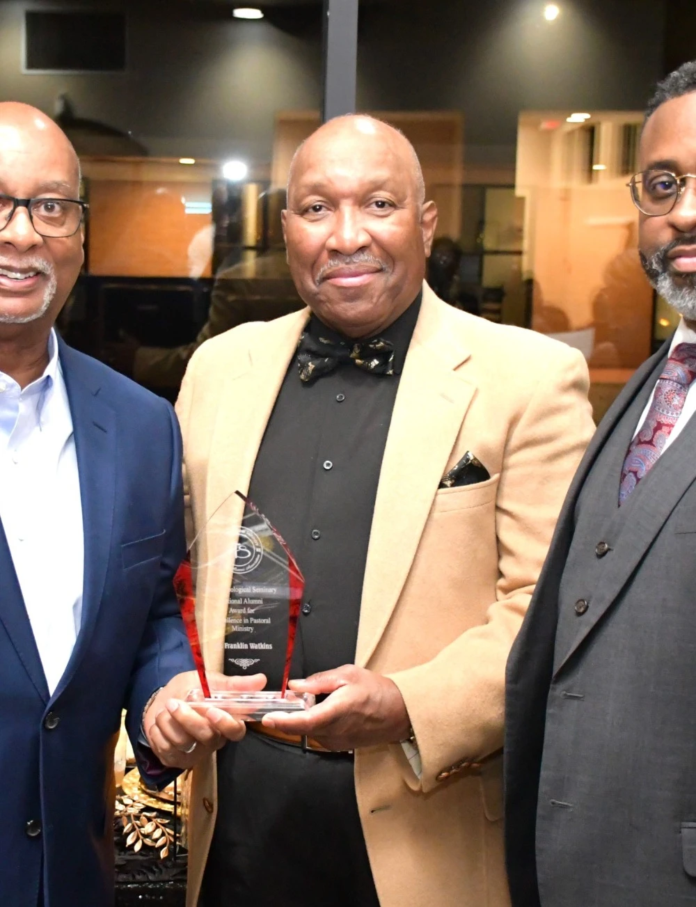 An alumni award recipient in a tan jacket holding his award with fellow attendees.